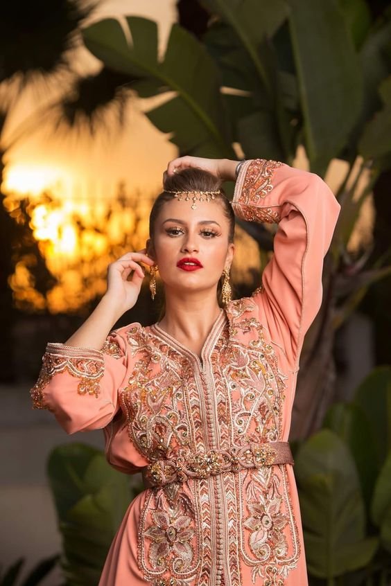 Caftan marocain traditionnel classique pour femme élégante, en satin de soie couleur pêche, orné de broderies dorées complexes et de perles, porté lors d'une soirée en extérieur.